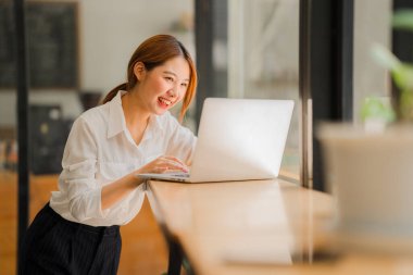 Çekici Asyalı kadın kafede bir masada tek başına oturuyor iş için dizüstü bilgisayarına bakıyor gibi görünüyor ve gülümsüyor, internette çalışıyor gibi..