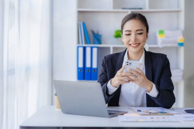 Asyalı iş kadını müşterilerle iletişim kurmak için akıllı telefon ve dizüstü bilgisayar kullanıyor, masadaki hesap makinesiyle hesaplıyor, finansal grafikleri okuyor, planlıyor, analiz ediyor, şirket ofisindeki pazar verilerini hesaplıyor..