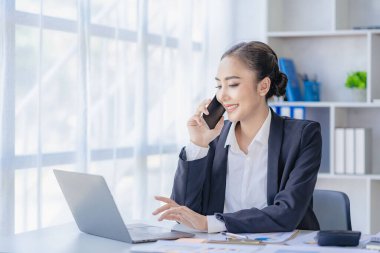 Asyalı iş kadını müşterilerle iletişim kurmak için akıllı telefon ve dizüstü bilgisayar kullanıyor, masadaki hesap makinesiyle hesaplıyor, finansal grafikleri okuyor, planlıyor, analiz ediyor, şirket ofisindeki pazar verilerini hesaplıyor..