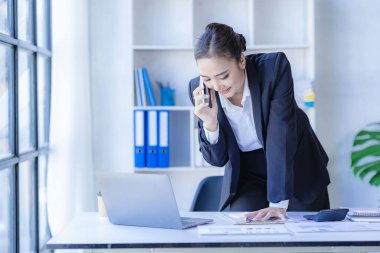 Asyalı iş kadını müşterilerle iletişim kurmak için akıllı telefon ve dizüstü bilgisayar kullanıyor, masadaki hesap makinesiyle hesaplıyor, finansal grafikleri okuyor, planlıyor, analiz ediyor, şirket ofisindeki pazar verilerini hesaplıyor..