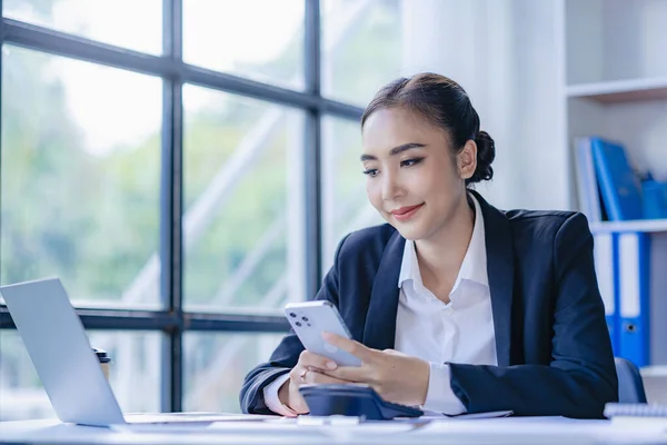 stock image Asian businesswoman using smartphone and laptop to contact customers, calculate with calculator on desk, reading financial graphs, planning, analyzing, calculating market data in corporate office.