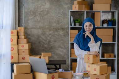 Genç müslüman kadın tesettürlü evde kutu ve dizüstü bilgisayarla çalışıyor çevrimiçi alışveriş sitelerinde müşterilere teslimat için kontrol paketleri hazırlıyor..