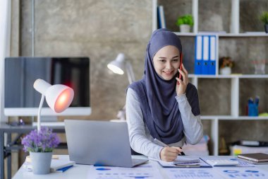 İşteki tesettürlü mutlu Müslüman iş kadını laptopta çalışan gülümseyen Arap kadın ve akıllı telefonla konuşan masada para grafiği, finansal muhasebe konsepti