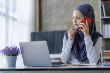 İşteki tesettürlü mutlu Müslüman iş kadını laptopta çalışan gülümseyen Arap kadın ve akıllı telefonla konuşan masada para grafiği, finansal muhasebe konsepti