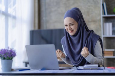 Heyecanlı Müslüman kadın portresi evde dizüstü bilgisayarla başarıyı kutluyor. Çok mutlu Asyalı kadın bilgisayar ekranına bakıyor Yumruğunuzu kaldırın, finansal iş kavramı