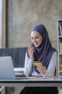 Mutlu genç Müslüman kadın tesettürlü, tesettürlü, elinde bankamatik kartı olan ve dizüstü bilgisayarından sipariş veren ofis alışverişi yapan banka hesabında internet üzerinden işlem yapan.