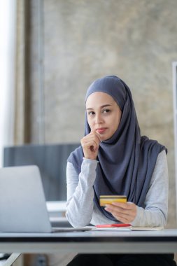 Mutlu genç Müslüman kadın tesettürlü, tesettürlü, elinde bankamatik kartı olan ve dizüstü bilgisayarından sipariş veren ofis alışverişi yapan banka hesabında internet üzerinden işlem yapan.
