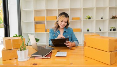 Çevrimiçi alışveriş kavramı, genç bayan girişimci, mutlu Asyalı KOBİ işletme sahibi paket kutuları hazırlıyor ve dizüstü bilgisayardaki müşterilere teslim etmek üzere çevrimiçi ürün siparişlerini kontrol ediyor.