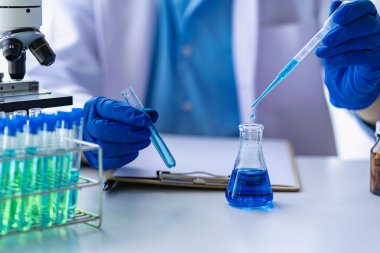 Doktor mavi sıvıyla test tüpü alıyor. Virolog, kimyasal koruyucu giysi üzerinde çalışıyor. Laboratuvar masasında tehlikeli virüs biyoloğu üzerinde çalışıyor.