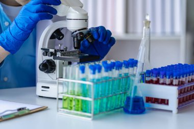 Doktor mavi sıvıyla test tüpü alıyor. Virolog, kimyasal koruyucu giysi üzerinde çalışıyor. Laboratuvar masasında tehlikeli virüs biyoloğu üzerinde çalışıyor.
