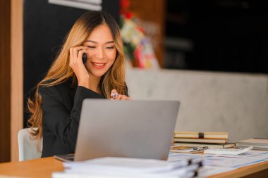 Asyalı iş kadını, bilgisayarlı ve mali raporlu akıllı telefon muhasebe defteri kullanıyor. Ofisteki ahşap masada matematik muhasebesi yapıyor. Vergi, muhasebe, finans kavramı.