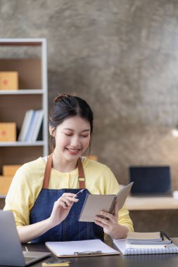 Dağıtım konsepti KOBİ, serbest çalışan Asyalı kadın dizüstü bilgisayarla gülerek kutulanmış ürünleri, çevrimiçi pazarlamayı ve evde satış yapmayı başarıyor..