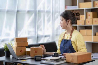 Dağıtım konsepti KOBİ, serbest çalışan Asyalı kadın dizüstü bilgisayarla gülerek kutulanmış ürünleri, çevrimiçi pazarlamayı ve evde satış yapmayı başarıyor..