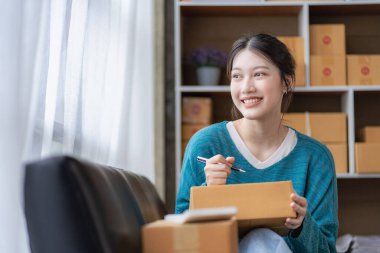 Dağıtım konsepti KOBİ, serbest çalışan Asyalı kadın dizüstü bilgisayarla gülerek kutulanmış ürünleri, çevrimiçi pazarlamayı ve evde satış yapmayı başarıyor..