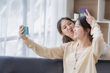 Genç ve güzel Asyalı lezbiyen mutlu çift evdeki oturma odasında akıllı telefon kullanarak kanepede oturuyor. LGBT lezbiyen çifti birlikte yaşıyor.