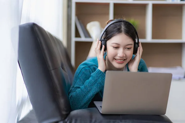 Young Asian woman listening to music on the sofa in the living room at home. Happy woman using laptop to video call with friends on vacation