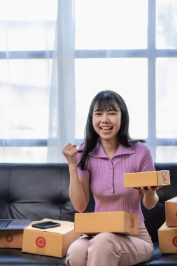 Kanepede oturan ve evde çalışan Asyalı kadın, posta kutusuna internet üzerinden satış ürünleri göndermek için müşteri adresi yazmak için heyecanlı ve gülümsüyor. KOBİ işletme sahipleri kavramı