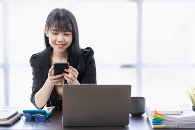 Bilgisayarla çalışan Asyalı iş kadını iş arkadaşları ve müşterilerle iletişim kurmak için cep telefonu kullanıyor. Finansal konsept ofisinde modern teknolojiyi kullanarak çalıştır ve koordine et.