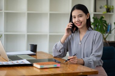 Bilgisayarla çalışan Asyalı iş kadını iş arkadaşları ve müşterilerle iletişim kurmak için cep telefonu kullanıyor. Finansal konsept ofisinde modern teknolojiyi kullanarak çalıştır ve koordine et.