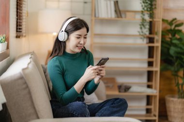 Oturma odasındaki kanepede oturup boş günlerinde dinlenen kulaklık takan mutlu Asyalı kadın.