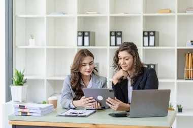 Beyaz tenli genç patron ve asistanı, ofis masasındaki grafik kağıdı ve tablet dizüstü bilgisayara danışmanlık yapıyor.