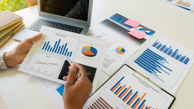 Finans müdürünün elleri masadaki, iş planındaki raporları analiz ediyor ve birçok grafiği dizüstü bilgisayar, hesap makinesi, kalem, üst görünüm ile analiz ediyor..