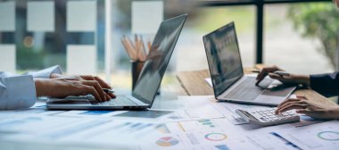 İş arkadaşı kağıt belgeleri, hesaplar, sözleşmeler, yatırım danışmanı, dizüstü bilgisayar ve analitik teknoloji kullanarak kurumsal pazarlama planları üzerinde çalışıyor ve analiz ediyor..