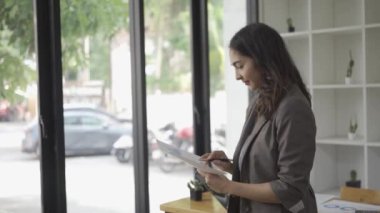 Masanın önünde duran güzel, uzun saçlı Asyalı iş kadını iyi giyimli, başarılı bir kadın ofiste kendine güvenen bir gülümsemeyle kameraya bakıyor.