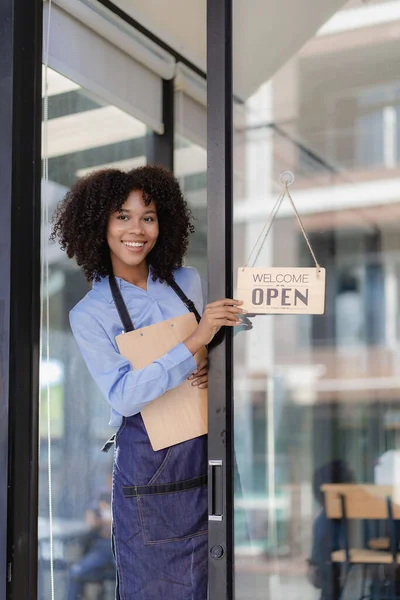 Young American woman standing in front of the restaurant door with an open sign. She is a waitress at a restaurant and coffee shop preparing to open. to serve customers food and beverage concept