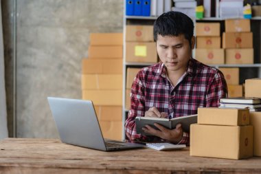 Kareli gömlekli genç bir Asyalı işadamı ev ofisinde dizüstü bilgisayarı ve kutusuyla çalışıyor. KOBİ 'nin online pazarlama için bir şirket kurma konsepti gibi görünüyor. Paket teslim kavramı.
