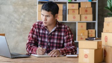 Kareli gömlekli genç bir Asyalı işadamı ev ofisinde dizüstü bilgisayarı ve kutusuyla çalışıyor. KOBİ 'nin online pazarlama için bir şirket kurma konsepti gibi görünüyor. Paket teslim kavramı.