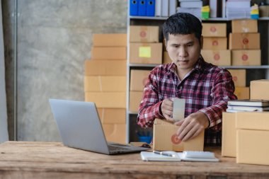 Kareli gömlekli genç bir Asyalı işadamı ev ofisinde dizüstü bilgisayarı ve kutusuyla çalışıyor. KOBİ 'nin online pazarlama için bir şirket kurma konsepti gibi görünüyor. Paket teslim kavramı.