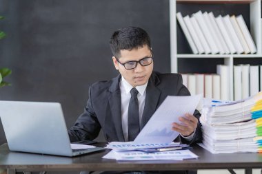 Asyalı finansör, evrak ve dizüstü bilgisayarla muhasebe işlerini yapıyor. İşadamı, ofis binasında yığınla evrakla masada oturuyor.