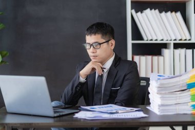 Asyalı finansör, evrak ve dizüstü bilgisayarla muhasebe işlerini yapıyor. İşadamı, ofis binasında yığınla evrakla masada oturuyor.