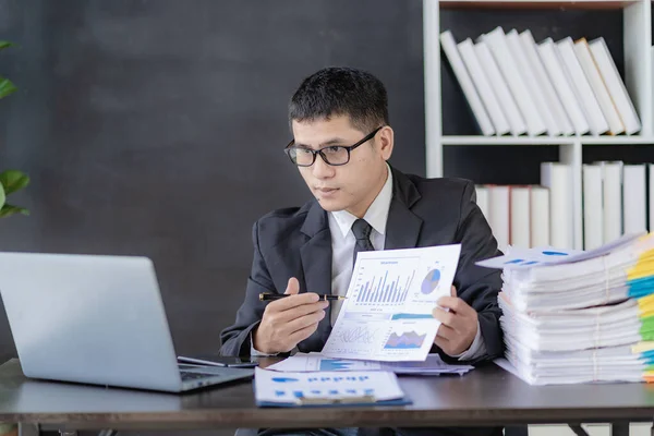 Asian financier doing accounting work in glasses working with documents and laptop Businessman sitting at desk with stacks of papers in office building