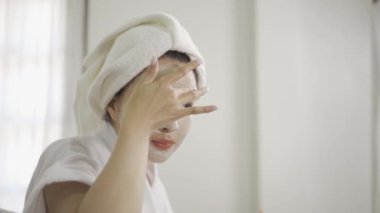 Beautiful young Asian woman doing make-up at home with towel on her head is wearing mask at home.