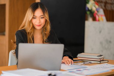 Asian businesswoman working on charts and graphs showing results in office with documents and laptop Worker documents calculating financial indicators, smiling and happy with success.