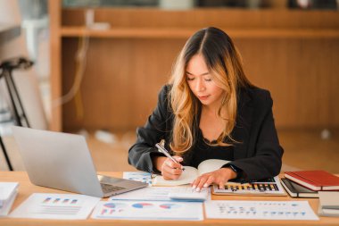 Asian businesswoman working on charts and graphs showing results in office with documents and laptop Worker documents calculating financial indicators, smiling and happy with success.