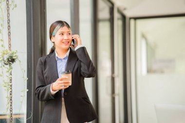 Attractive asian businesswoman or accountant holding cup of coffee and talking on the phone with her business clients while working in the office vertical image