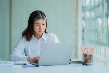 Asian female accountant doing calculations analyzing reports pointing at graphs holding pen using laptop placed on table at office