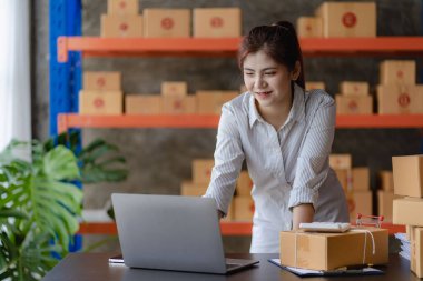Young Asian woman checking parcels for delivery taking orders with small business laptop. SME business owners check orders online Prepare to pack products for sale to customers, SME business ideas