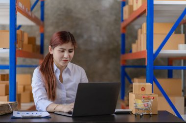 Young Asian woman checking parcels for delivery taking orders with small business laptop. SME business owners check orders online Prepare to pack products for sale to customers, SME business ideas