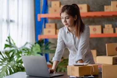 Young Asian woman checking parcels for delivery taking orders with small business laptop. SME business owners check orders online Prepare to pack products for sale to customers, SME business ideas