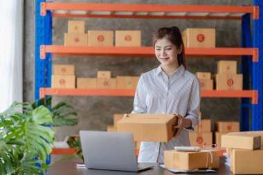 Young Asian woman checking parcels for delivery taking orders with small business laptop. SME business owners check orders online Prepare to pack products for sale to customers, SME business ideas