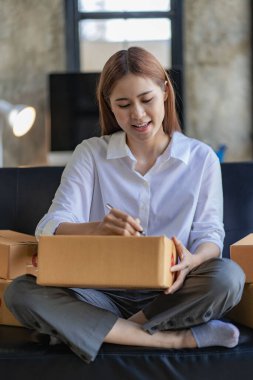 Happy Asian young woman business owner preparing parcel boxes and checking orders for delivery to customer on laptop computer at home, SME marketing, online shopping concept.