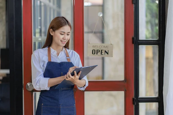 Asian woman in apron holding restaurant menu in front of door with open sign, small restaurant concept