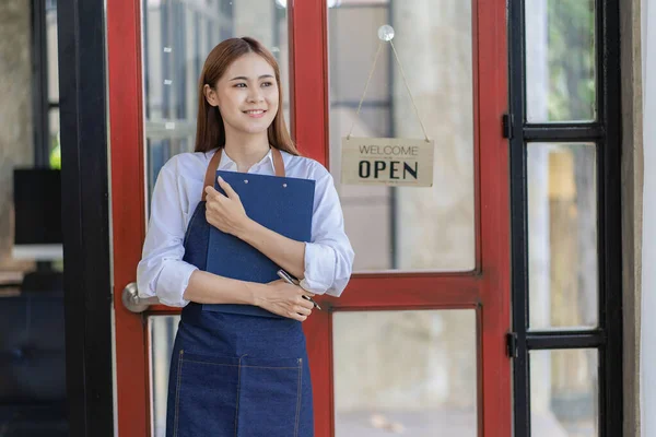 Asian woman in apron holding restaurant menu in front of door with open sign, small restaurant concept