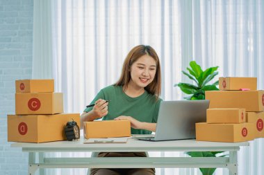 SME business concept, online shopping, happy asian female business owner prepares parcel boxes and checks online orders of products for delivery to customers on laptop computer.