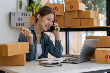 SME business concept, online shopping, happy asian female business owner prepares parcel boxes and checks online orders of products for delivery to customers on laptop computer.