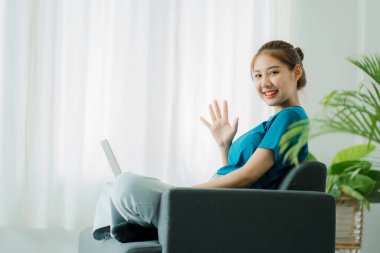 Young beautiful and cheerful asian woman in casual clothes talking with friends on video call on laptop using social media while relaxing in living room on sofa at home.